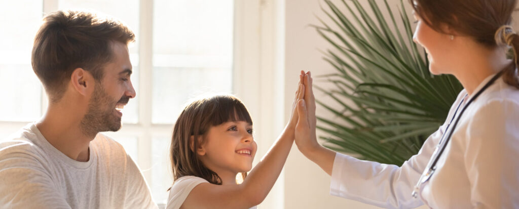 A pediatrician giving a high-five to a child while a parent smiles nearby during a well-child check-up.