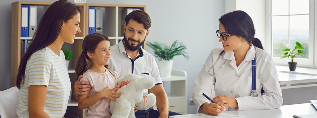 A smiling family—mother, father, and young daughter—sits with a female doctor in a bright, modern medical office. The child holds a white stuffed animal while the doctor takes notes.