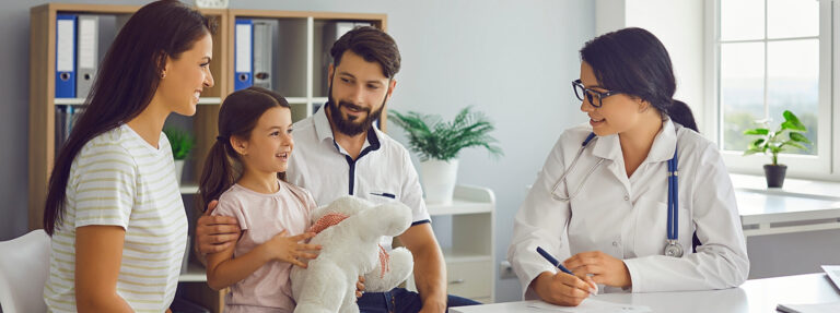 A smiling family—mother, father, and young daughter—sits with a female doctor in a bright, modern medical office. The child holds a white stuffed animal while the doctor takes notes.