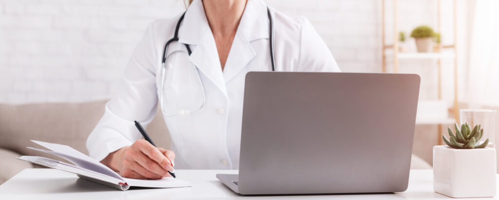Female doctor in a white coat writing in a notebook while working on a laptop at a clean, minimalist desk.
