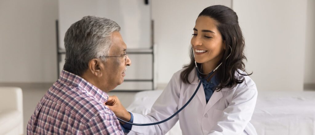 Doctor smiling while checking a senior patient’s heartbeat with a stethoscope during a chronic disease management visit.