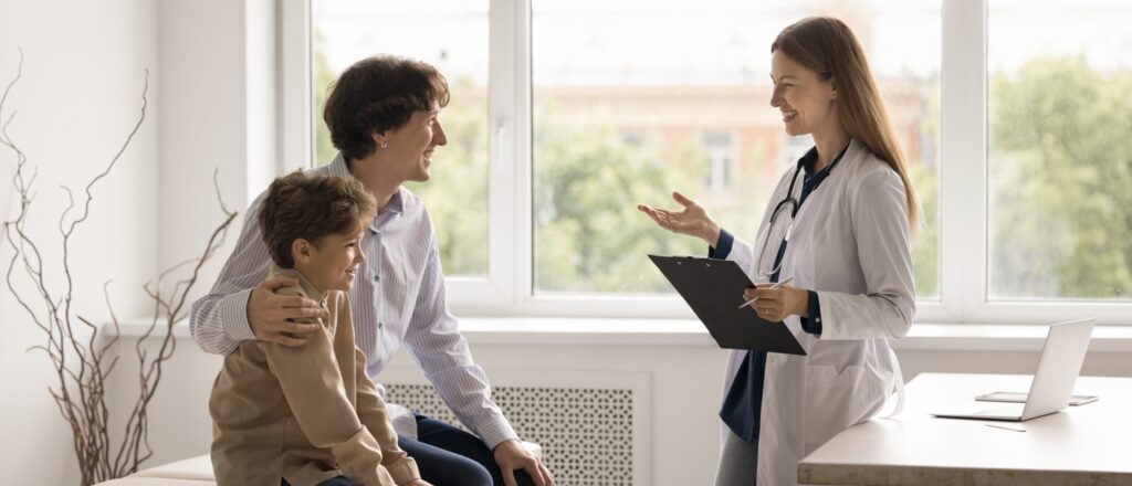 Smiling doctor talking to a father and child during a relaxed medical appointment in a bright, welcoming clinic.