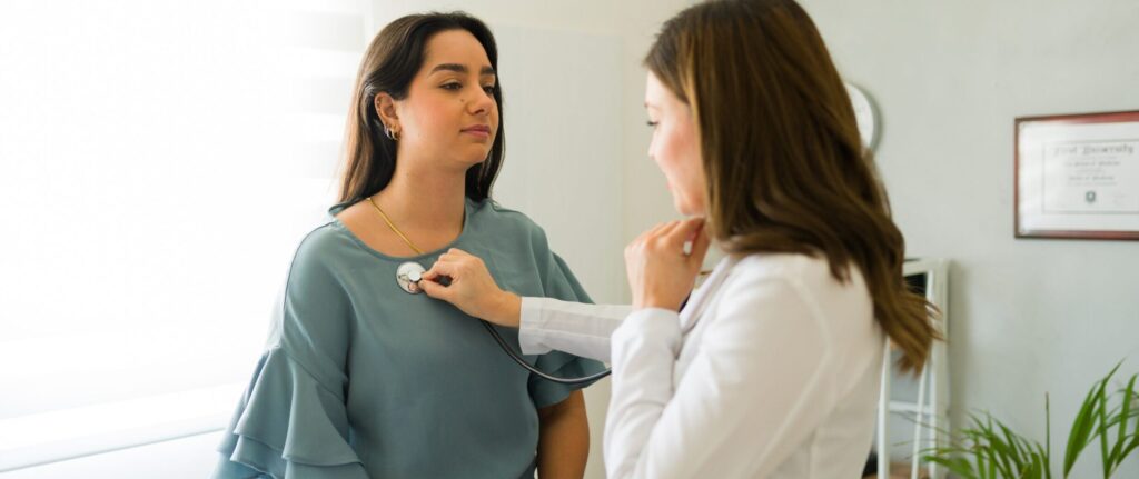 A female healthcare provider using a stethoscope to listen to a patient’s heartbeat during a routine checkup at a primary care office.